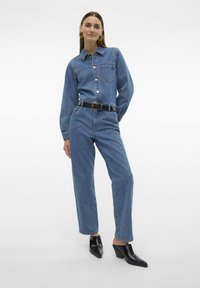 Macacão de ganga em azul claro, com mangas longas, frente com botões e um bolso no peito. Combinado com mules pretos de salto e um cinto preto.
