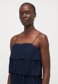 Navy blue pleated dress with layered ruffles, thin spaghetti straps, and a smooth texture. Simple design with a flowing silhouette.