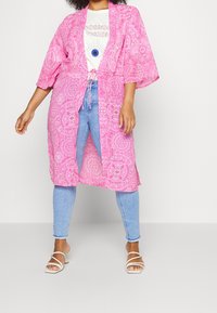 Modell iklädd en rosa m önstrad kimonojacka över en vit t-shirt med grafik och ljusblå jeans, ihop med vita sandaler.