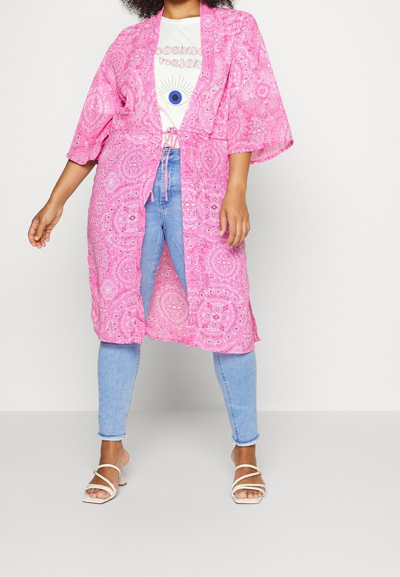 Modell iklädd en rosa mönstrad kimonojacka över en vit t-shirt med grafik och ljusblå jeans, ihop med vita sandaler.