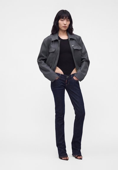 Jeune femme aux longs cheveux foncés portant une veste grise, un body noir, un jean foncé et des sandales noires, debout les mains dans les poches.