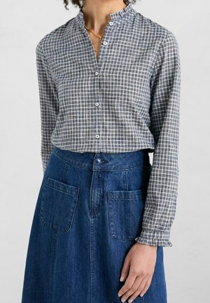 Vrouw draagt een grijs geruit hemd met lange mouwen, een gerimpelde kraag en manchetten, ingestopt in een hooggetailleerde blauwe denimrok met voorzakken.