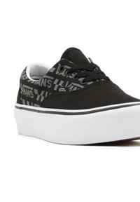 Black canvas sneaker with white rubber sole, featuring a repeated "VANS" logo pattern and black laces. Smooth texture, low-top design.