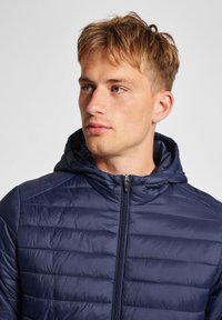 Giacca trapuntata blu navy con cappuccio, caratterizzata da una texture trapuntata, chiusura con zip e un design leggero. Finitura in tessuto liscio e lucido.