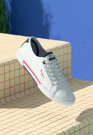 Białe trampki Pepe Jeans z sznurówkami oraz czerwonym i granatowym paskiem wzdłuż podeszwy, umieszczone na beżowych, wyłożonych kafelkami schodach na tle niebieskiej ściany.