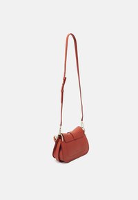 Petit sac bandoulière en cuir rouge avec sangle réglable et quincaillerie dorée, présenté suspendu sur fond blanc.