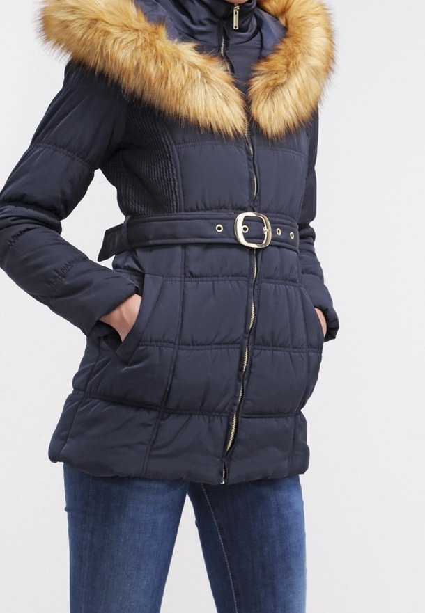 Femme portant une veste d'hiver matelassée bleu marine avec une capuche bordée de fourrure marron, taille cintrée par une ceinture, les mains dans les poches, associée à un jean bleu.