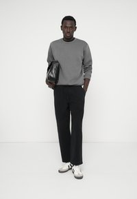 Szürke hosszú ujjú pulóver, fekete bővebb szabású nadrág és fekete részletekkel ellátott fehér sportcipők. Fekete táskát tart a kezében. Egyszerű és modern megjelenés.