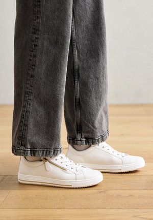 Person iført grå vide jeans og hvide lædersneakers med lynlåse på siden, stående på trægulv.