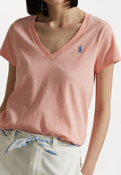 Pink cotton t-shirt with a v-neck, short sleeves, and blue embroidered logo on the left chest. Worn with white pants and a floral belt.
