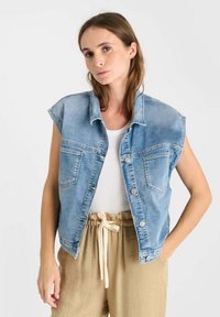 Blauwe denim vest met korte mouwen, twee borstzakken en een knoopsluiting. Draagt over een witte top met beige drawstring-broek.