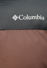 Logo Columbia v bielej farbe na dvojtonovej bunde so svetlo šedou vrchnou a červenohnedou spodnou časťou s jemnou mriežkovou štruktúrou.