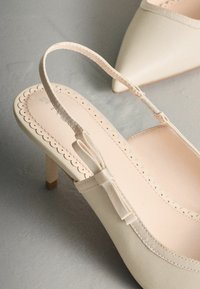 Smėlio spalvos bateliai su dirželiais ant kulno, smailiu pirštu, banguotais kraštais ir plonu, tekstūruotu dirželiu; turi mažą bloko formos kulną.