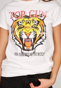 Hvid t-shirt med et slidt grafisk tryk af en brølende tiger ansigt og rød tekst "TOP GUN" ovenfor, båret af en person med hånden delvist i lommen på sorte bukser.