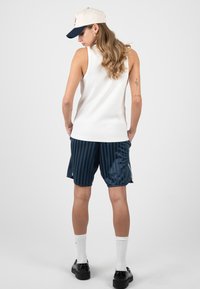 Femme portant un débardeur blanc, un short rayé bleu marine, une casquette blanche, des chaussettes blanches et des chaussures noires, debout les mains dans les poches, de dos.