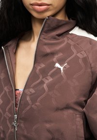 Κλείσιμο εικόνας ενός ατόμου που φοράει καφέ μπουφάν με φερμουάρ, με λογότυπο Puma σε λευκό και κυματιστό σχέδιο, εν μέρει φερμουάρ πάνω από πολύχρωμο τοπ.