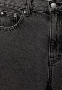 Gros plan sur un jean en denim noir montrant la poche avant, la petite poche, les rivets métalliques, la passant de ceinture et la fermeture par bouton avec des coutures visibles.