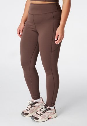 Vrouw die bruine high-waist legging met zijzak en roze, witte en zwarte sportschoenen draagt, staand tegen een witte achtergrond.