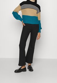 Frau trägt einen farblichen Blockstrickpullover und schwarze ausgestellte Hosen, steht mit einer Hand auf der Hüfte und schwarzen Slippern.