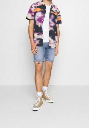Camisa de manga corta con un patrón tie-dye en púrpura, negro y naranja, combinada con pantalones cortos de mezclilla claros y zapatillas altas beige.