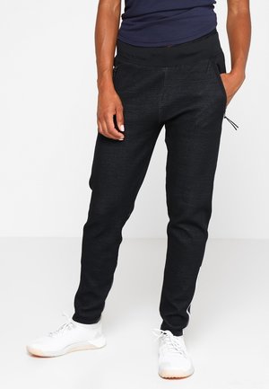 Persona con pantalones jogger negros texturizados con bolsillos laterales con cremallera y zapatos deportivos blancos, de pie contra un fondo liso.