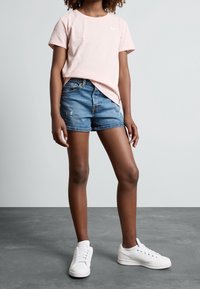 T-shirt di cotone rosa chiaro con maniche corte e un piccolo logo, abbinata a pantaloncini di jeans blu e sneaker bianche su sfondo grigio.