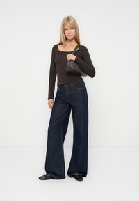 Langermet, mørkebrun topp med rund hals, kombinert med vide, mørkeblå jeans. Modellen holder en liten, grå håndveske. svarte sko.