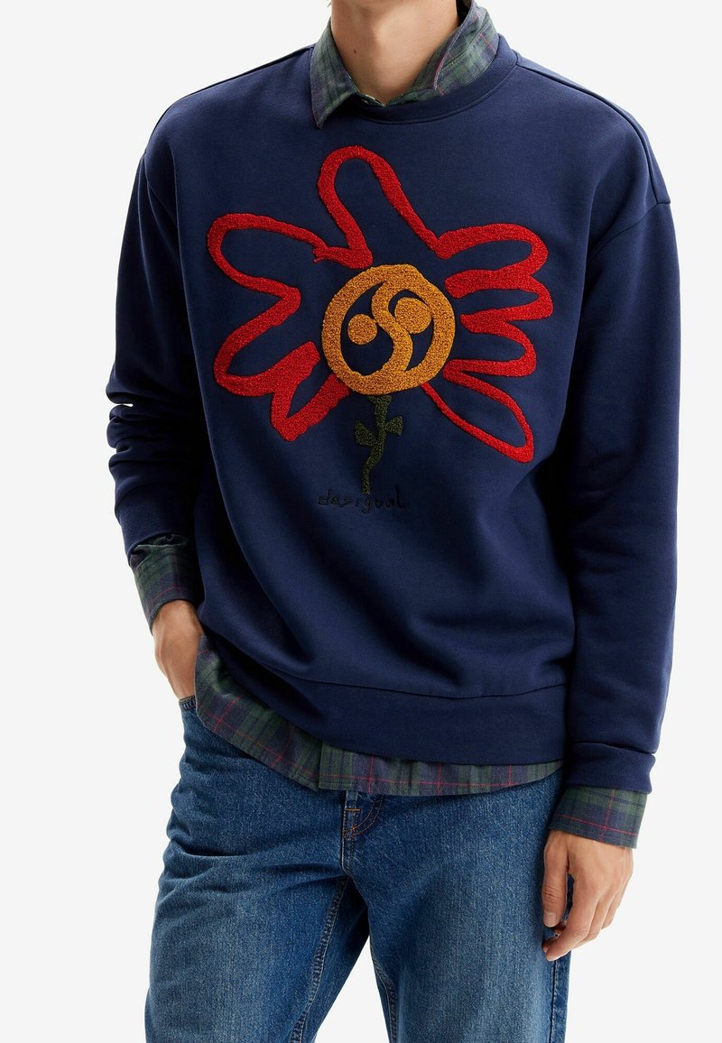 Homme portant un sweat-shirt bleu marine avec un motif floral abstrait rouge et orange, superposé sur une chemise à carreaux verte et un jean bleu.