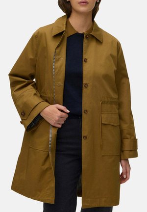 Femme portant un manteau marron boutonné par-dessus un haut bleu marine et un pantalon foncé, tenant le manteau légèrement ouvert d'une main.