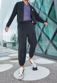 Chaqueta bomber de terciopelo negro sobre una camisa morada con cuello, combinada con pantalones negros entallados. Zapatillas coloridas con estampado y suelas blancas.