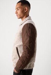 Giubbotto bomber texture con corpo beige chiaro, maniche imbottite marroni, cuciture a contrasto e tasche laterali. Vestibilità casual con polsini a coste.