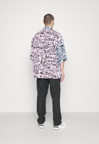 Camisa de manga corta en tela rosa y azul con texto gráfico negro y patrones. Llevada con pantalones negros y zapatillas blancas.