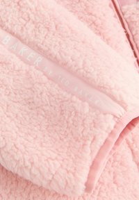 Veste en polaire rose avec un tissu doux et texturé. Elle dispose d'une fermeture éclair et d'une étiquette portant la mention "BAKER by TED BAKER" en blanc.