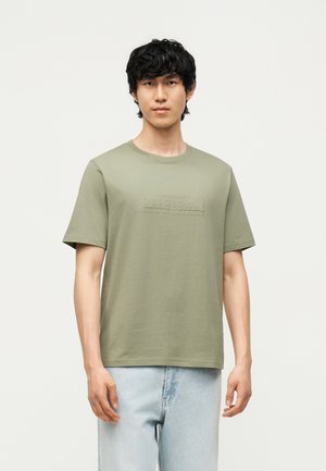Jeune homme aux cheveux noirs bouclés portant un t-shirt vert olive à manches courtes et un jean bleu clair sur un fond uni.