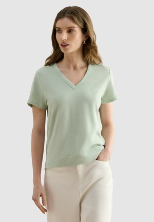 Mujer con cabello castaño claro que lleva una camisa de manga corta con escote en V de color verde claro y pantalones crema con una mano en el bolsillo sobre un fondo liso.