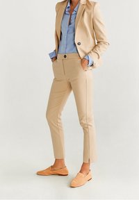 Kobieta ubrana w beżowy garnitur skrojony na miarę z krótkimi spodniami, niebieską koszulę na guziki i beżowe mokasyny, stojąca z rękoma w kieszeniach.