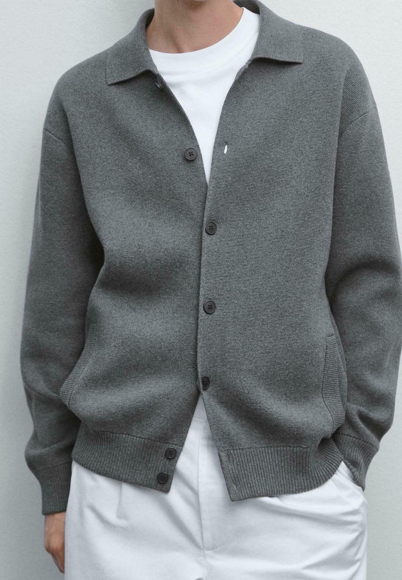 Personne portant un cardigan gris clair boutonné sur une chemise blanche et un pantalon blanc, debout les mains dans les poches sur un fond uni.
