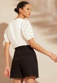 Bela bluzica z izrezljanimi naborki, v kombinaciji z visokimi črnimi hlačami, ki imajo valovit rob in zapleteno cvetlično vezenje.