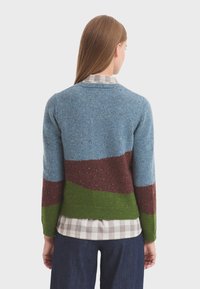 Pletený svetr s texturovanými modrými, hnědými a zelenými pruhy. Lehká příze se skvrnami. Nosený přes kostkovanou košili, ukazuje ležérní styl.