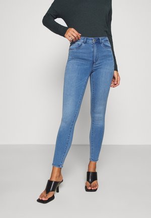 Vrouw draagt een hoge taille lichtblauwe skinny jeans, zwarte open-teensandalen met hakken en een lang mouwdonkergroene top, staand tegen een effen achtergrond.