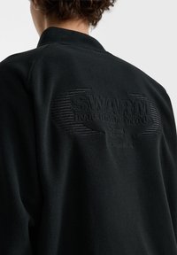Sort fleecejakke med et præg af logoet "SWARM INTERNATIONALS" på ryggen, med en tekstureret, blød overflade og minimalistisk design.