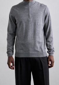 Pull en maille grise avec un col à demi-fermeture éclair, manches longues, poignets et ourlet côtelés, texture subtile, porté avec un pantalon noir.