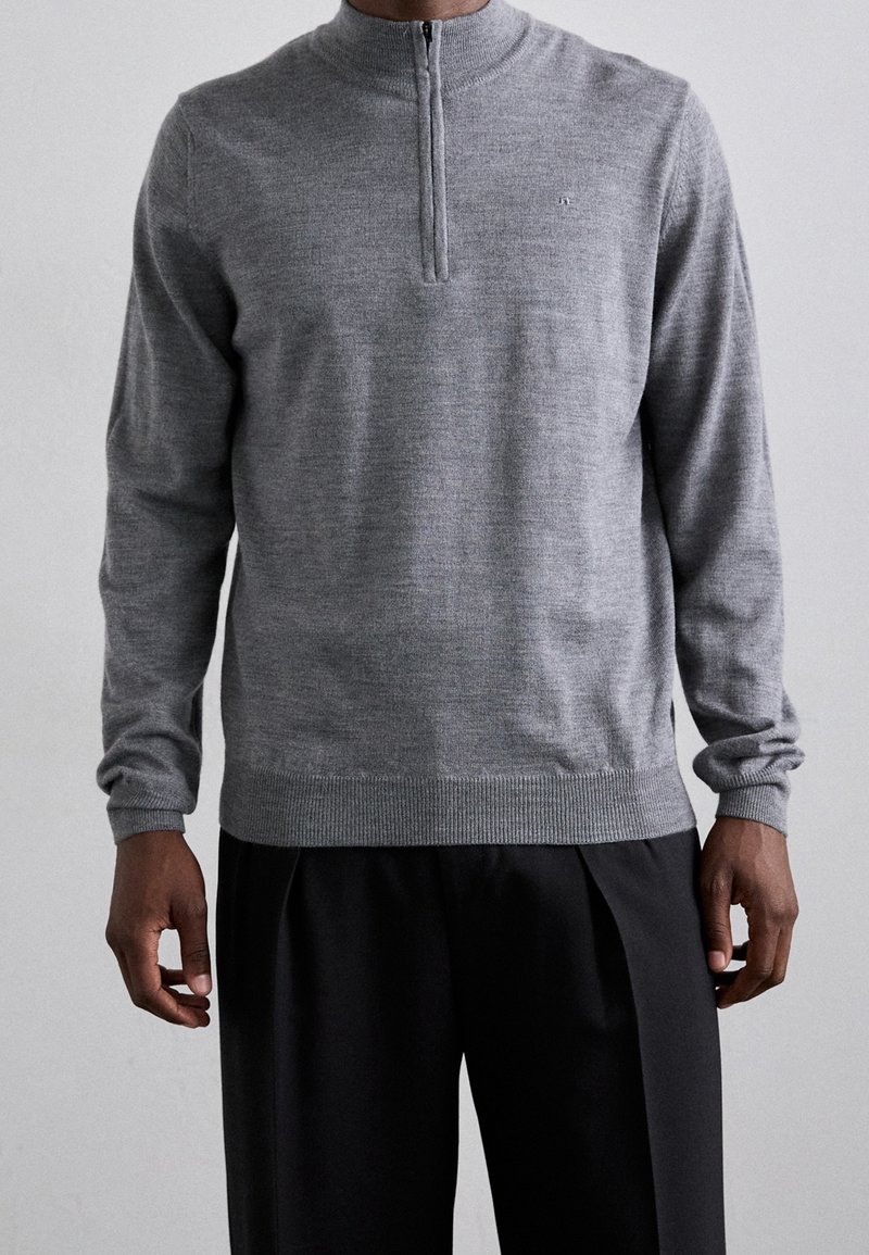 Pull en maille grise avec un col à demi-fermeture éclair, manches longues, poignets et ourlet côtelés, texture subtile, porté avec un pantalon noir.