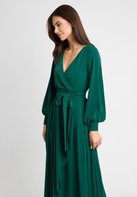 Zielona sukienka wrap z długimi, bufiastymi rękawami, dekoltem w serek i zawiązywaniem w talii. Wykonana z gładkiego materiału, z lejącą spódnicą.