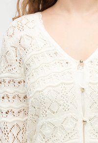 Moteris dėvi baltą nėrinių megztinį su širdies formos sagomis ir bangelėmis dekoruotais raštais, matomas viršutinis krūtinės ir pečių plotas.