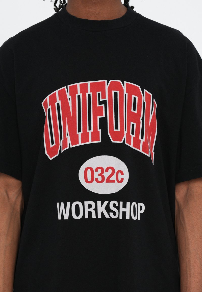 Sort bomulds t-shirt med fede røde og hvide grafikker, der står "UNIFORM 032c WORKSHOP". Korte ærmer og en klassisk rund halsudskæring.