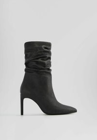 Botte en cuir noire mi-mollet décontractée avec bout pointu et talon stiletto haut sur un fond clair uni.