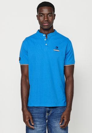 Polo blu con colletto henley, patta con tre bottoni, dettagli blu navy e un piccolo logo sul petto. Indossato con jeans in denim blu.