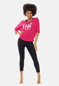 Leende kvinna med lockigt hår poserar barfota, iförd en rosa off-shoulder skjorta med texten "Först behöver jag kaffe" och svarta korta leggings.