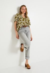 Blouse vert olive à motifs de feuilles ; jeans gris clair, légèrement courts ; sandales à talons carrés marron ; ceinture marron ; coupe décontractée et confortable.